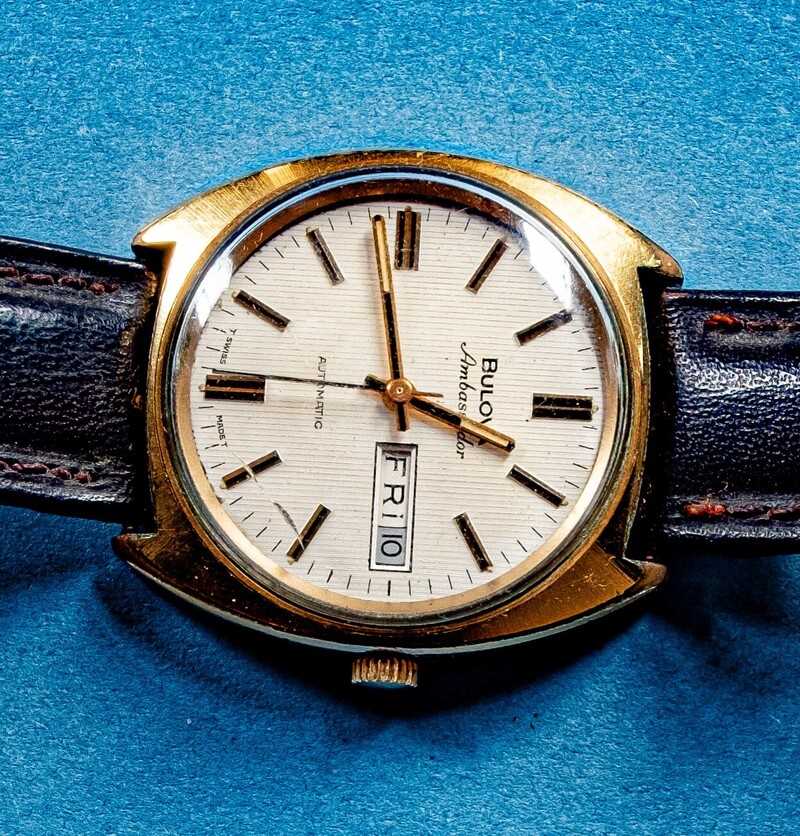 Dial of the watch showing the Bulova, Ambassador, 23 jewels, Automatic and Swiss Made info. Gold/cream dial with day and date and markers, not numbers, denote the time. Gold watch case, lugs and winder also shown.